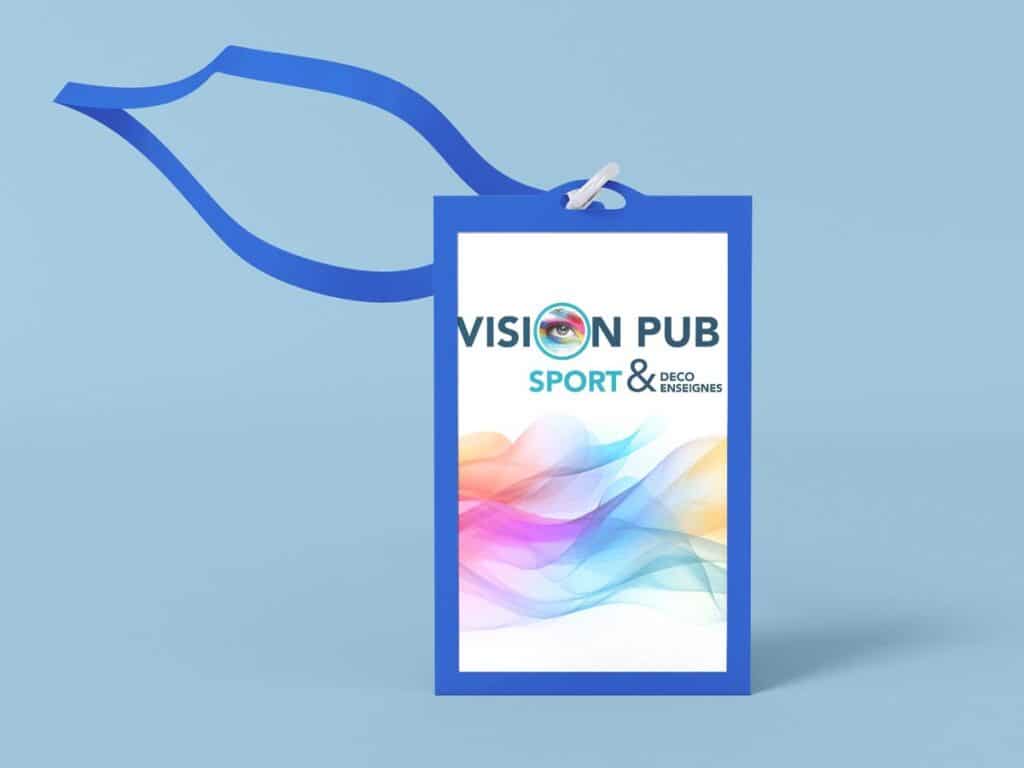 Badge bleu avec lanière bleue, affichant 'VISION PUB' avec un œil, et 'SPORT & DECO ENSEIGNES' sur un fond dégradé coloré.