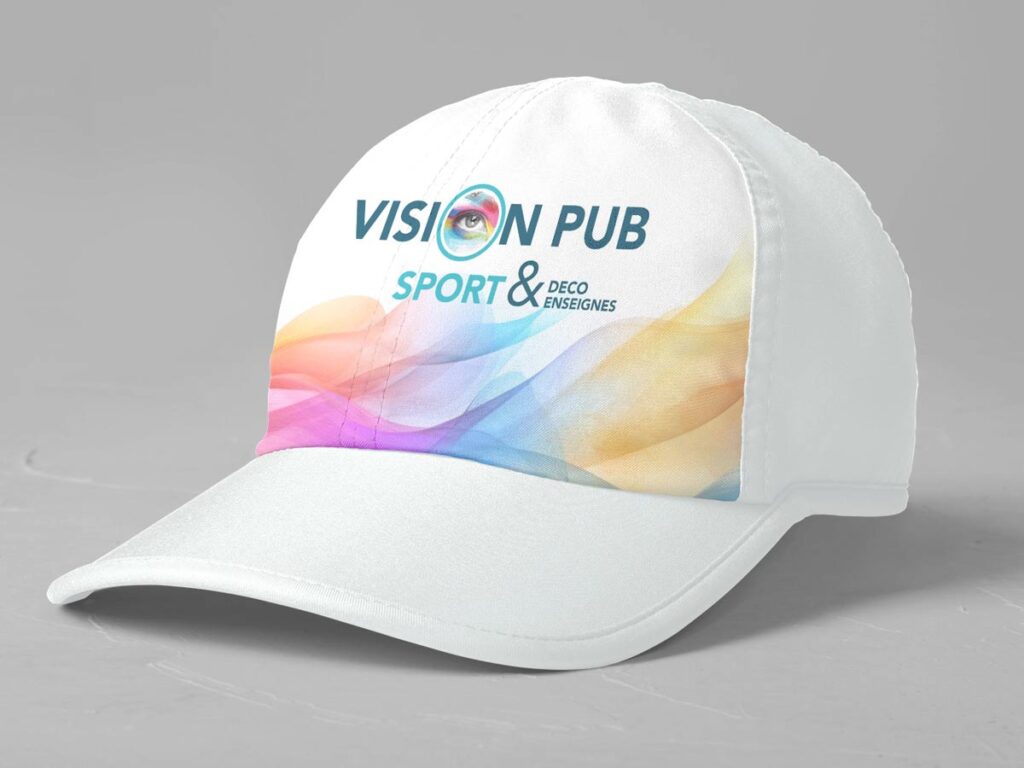 Casquette blanche personnalisée avec le logo 'VISION PUB' (œil stylisé) et un design abstrait coloré. Texte 'SPORT & DECO ENSEIGNES' visible.