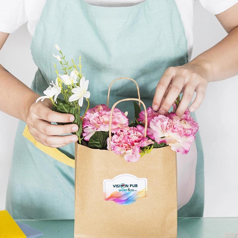 Personne en tablier arrangeant des fleurs roses et blanches dans un sac cadeau en papier kraft personnalisé avec sticker VISION PUB SPORT &.