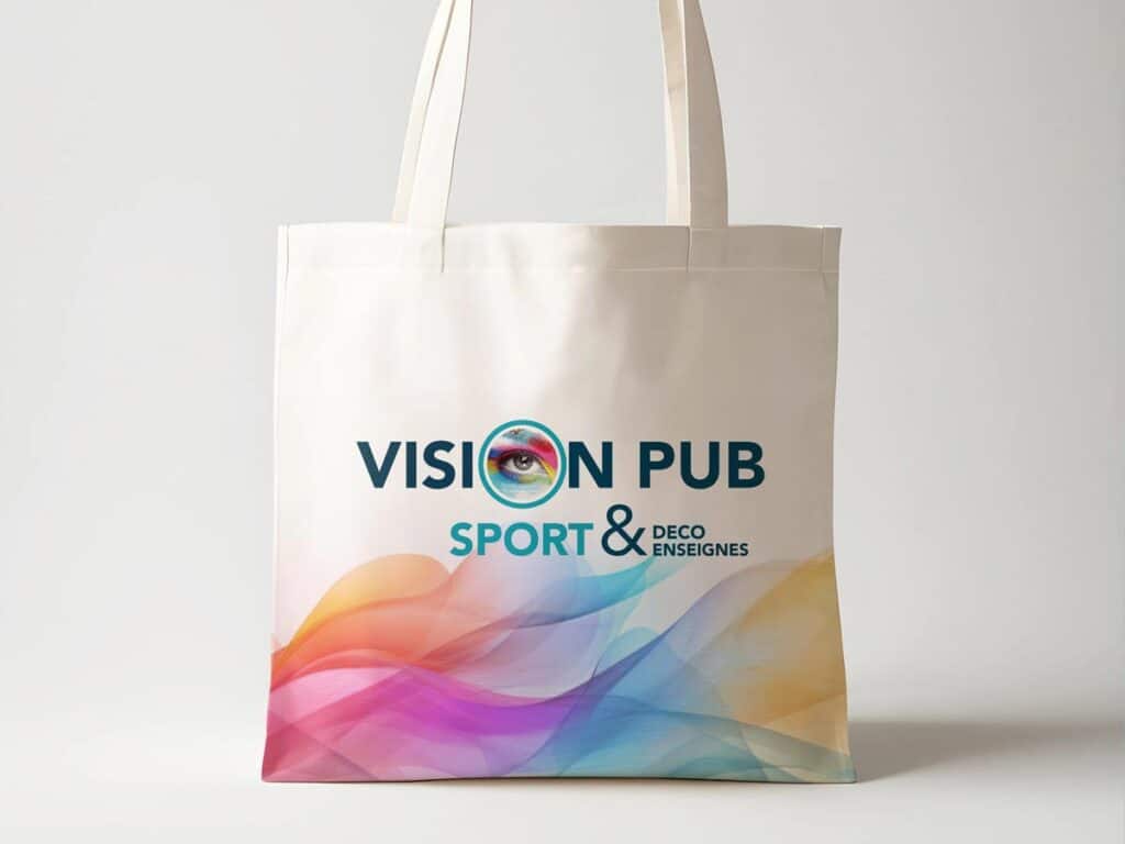 Sac tote bag blanc imprimé du logo "VISION PUB" avec un œil coloré, et le texte "SPORT & DECO ENSEIGNES". Un motif abstrait multicolore décore le bas.