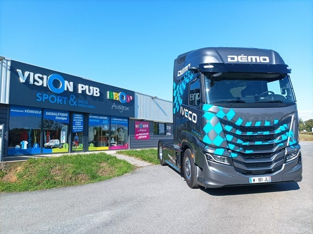 Camion Iveco DÉMO : Marquage Véhicule par Vision Pub Aveyron Camion Iveco gris avec motif à carreaux bleus 'DÉMO' devant un bâtiment 'VISION PUB BROD' Aveyron', spécialiste marquage véhicule, enseignes.