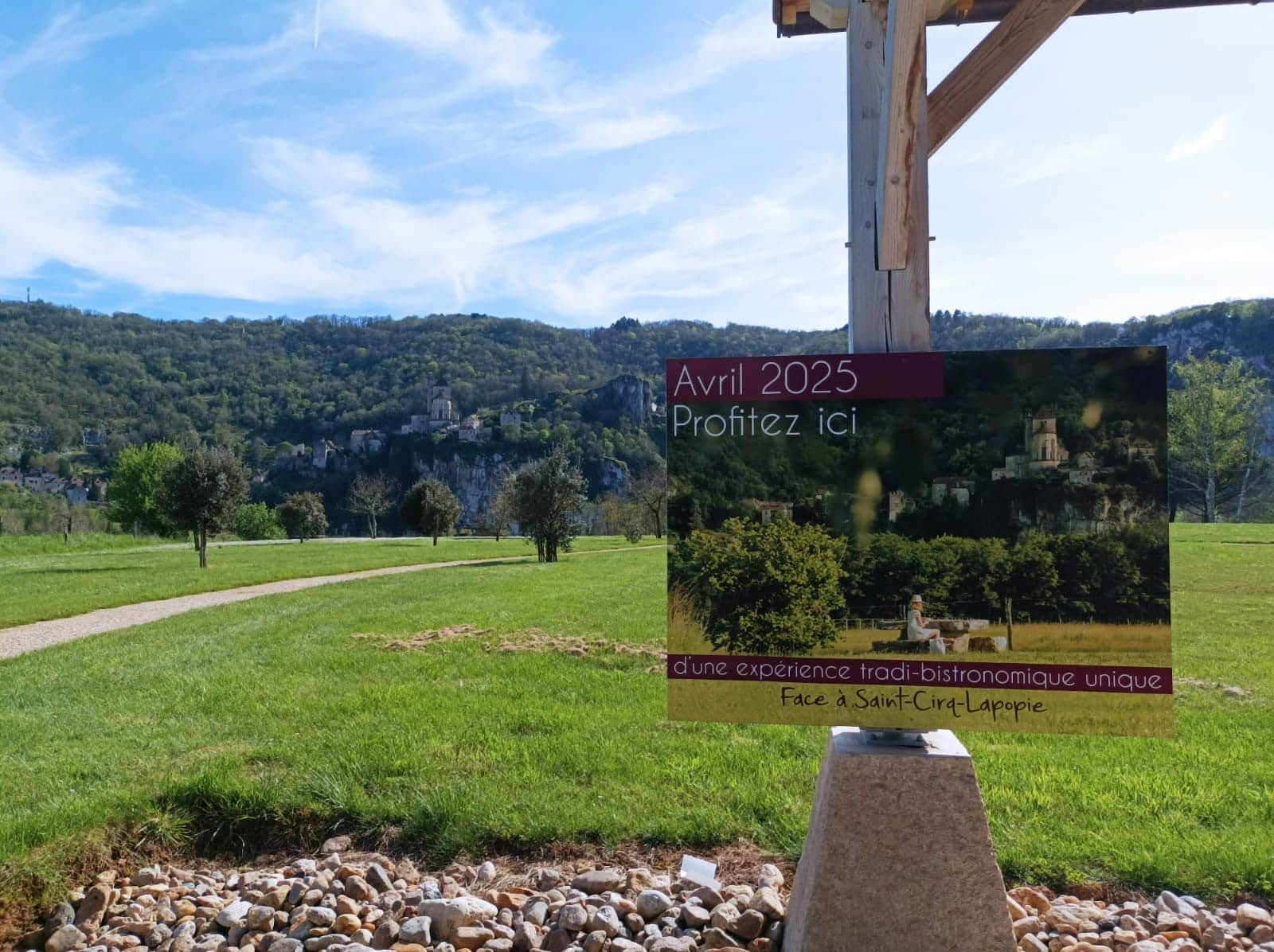 Champ verdoyant avec chemin face au village perché de Saint-Cirq-Lapopie. Un panneau annonce une expérience bistronomique unique dès Avril 2025.
