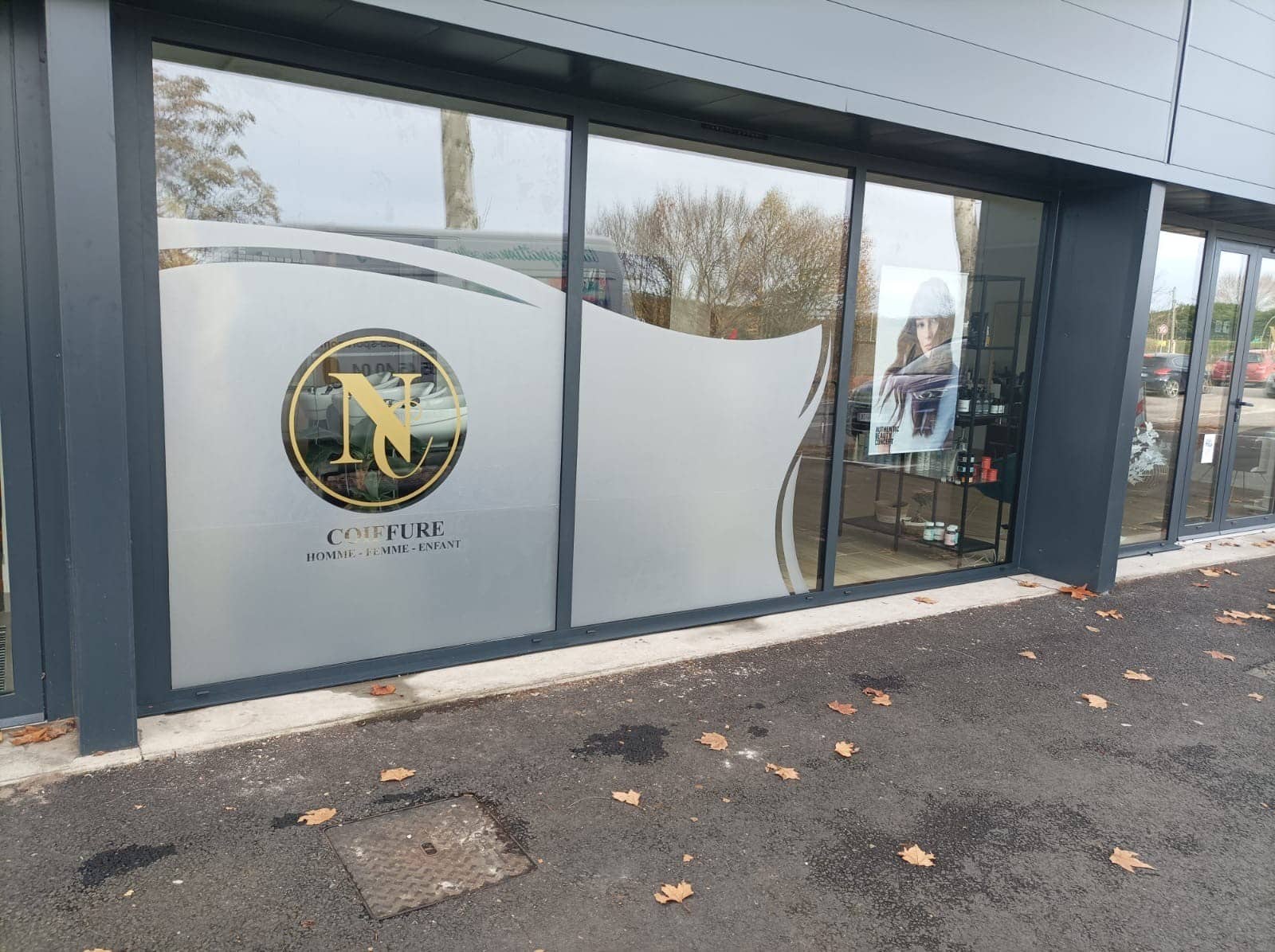 Façade vitrée d'un salon de coiffure avec bandes givrées, logo doré N&L, et l'inscription 'COIFFURE HOMME - FEMME - ENFANT'. Sol avec feuilles.
