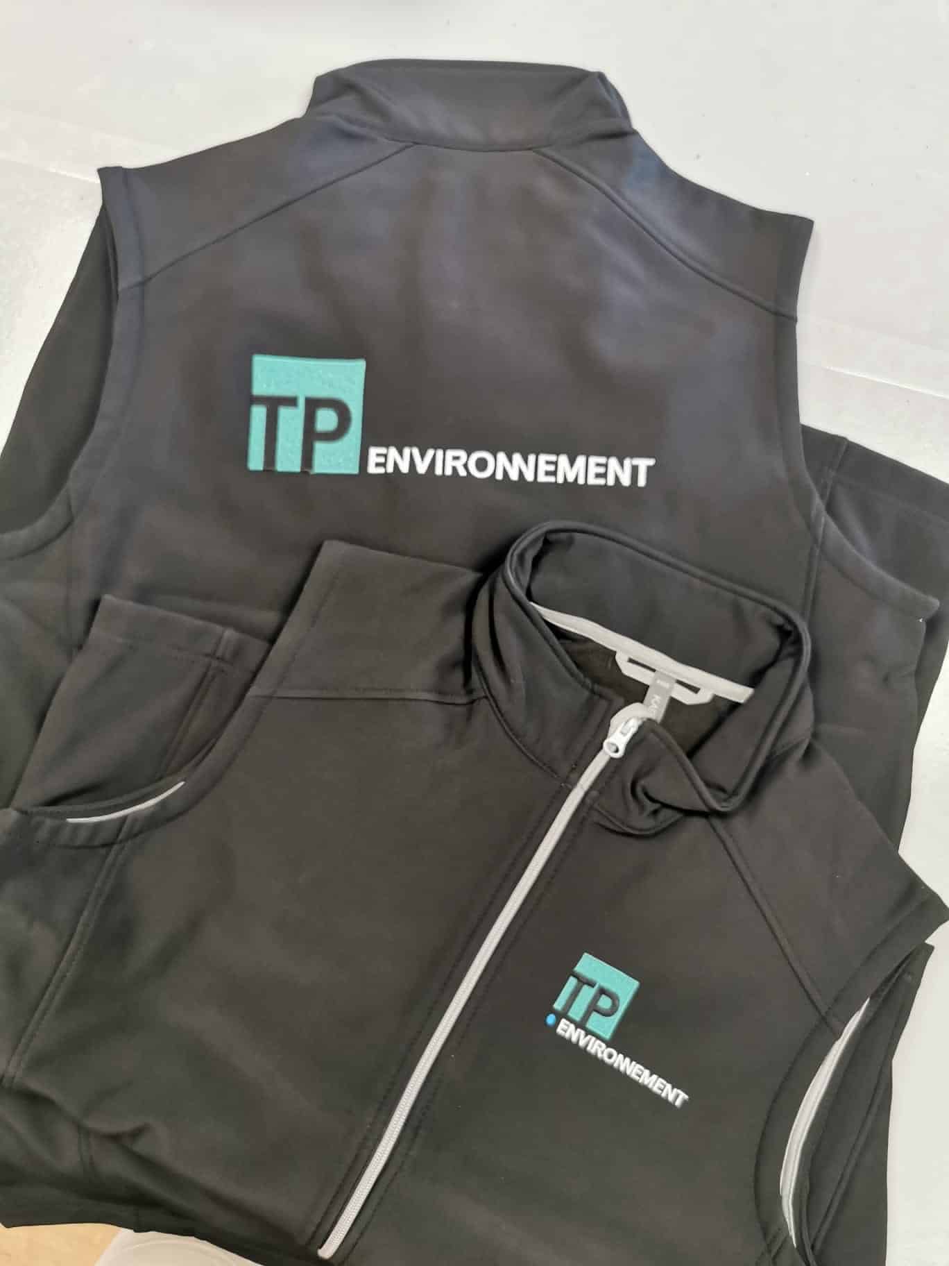 Vêtements professionnels noirs: une veste sans manches avec grand logo 'TP ENVIRONNEMENT' (turquoise/blanc) et une veste zippée avec petit logo.