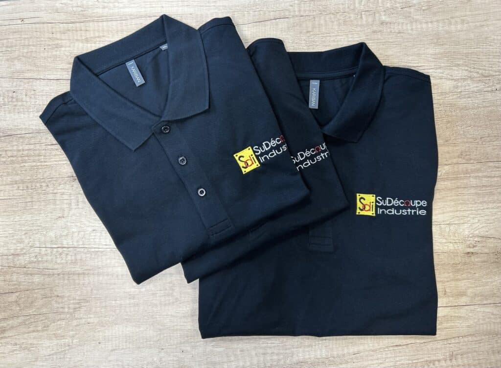 Polos Personnalisés SuDécoupe Industrie (SDI) Noirs Deux polos noirs Kariban marqués du logo brodé SuDécoupe Industrie (SDI) sur fond de bois clair.