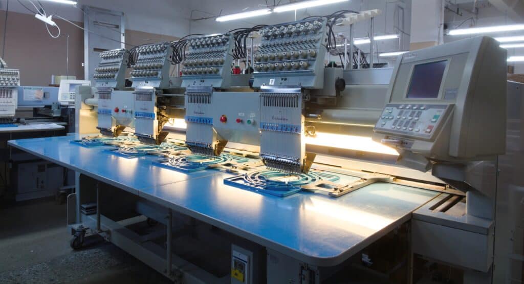 Machine à Broder Barudan Industrielle : Production Textile Machine à broder industrielle Barudan multicolore avec plusieurs têtes d'aiguilles, plateau bleu réfléchissant et panneau de commande lumineux.