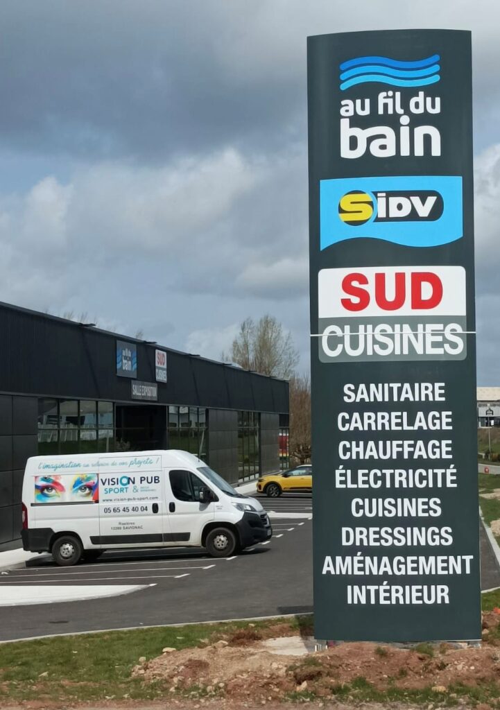 Enseignes Au fil du Bain, Sud Cuisines : Sanitaire & Aménagement Panneau signalétique d'un parc d'activités avec les enseignes Au fil du Bain, Sidv, Sud Cuisines et les services : Sanitaire, Carrelage, Électricité. Un van Vision Pub Sport est garé devant le bâtiment.