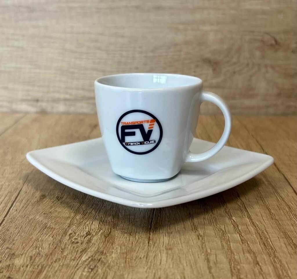 Tasse à café blanche et soucoupe carrée avec logo Transports FV Franck Viguie sur une table en bois.